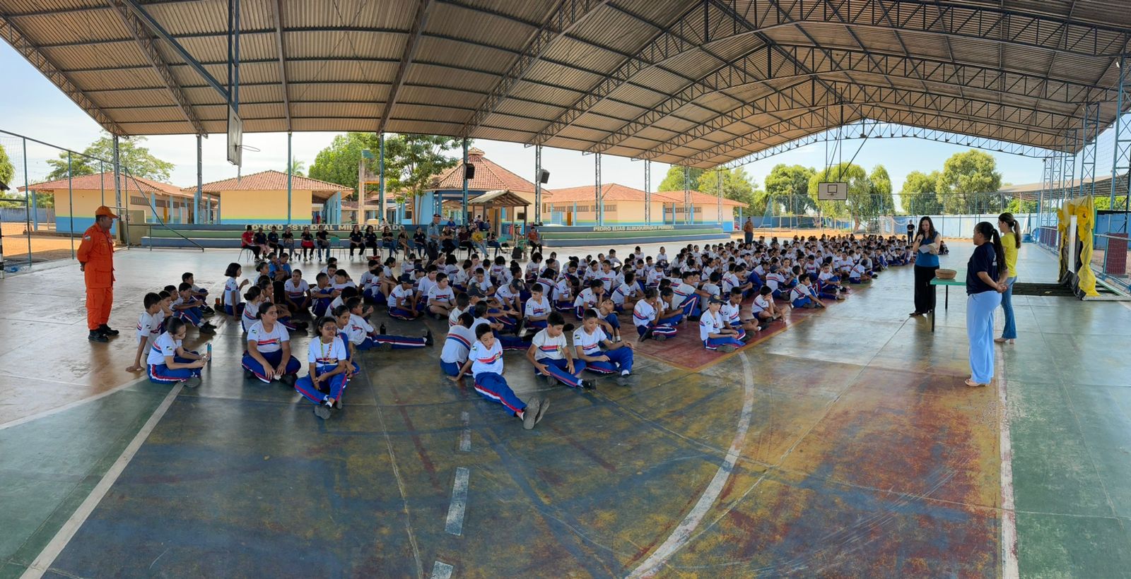 foto colorida de uma quadra escolar com vários estudantes sentados  no chão enquanto 3 mulheres então a frente e falando ao microfone