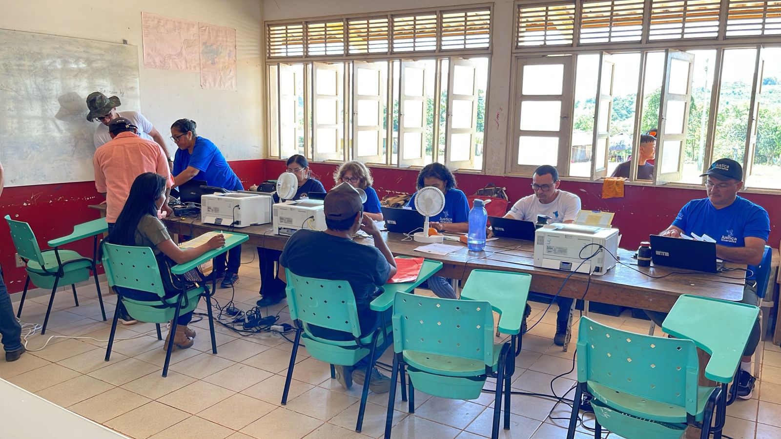 foto colorida de servidores da vara da justiça itinerante realizando atendimento dentro de uma sala. Há duas pessoas sentadas em mesas escolares de cor verde