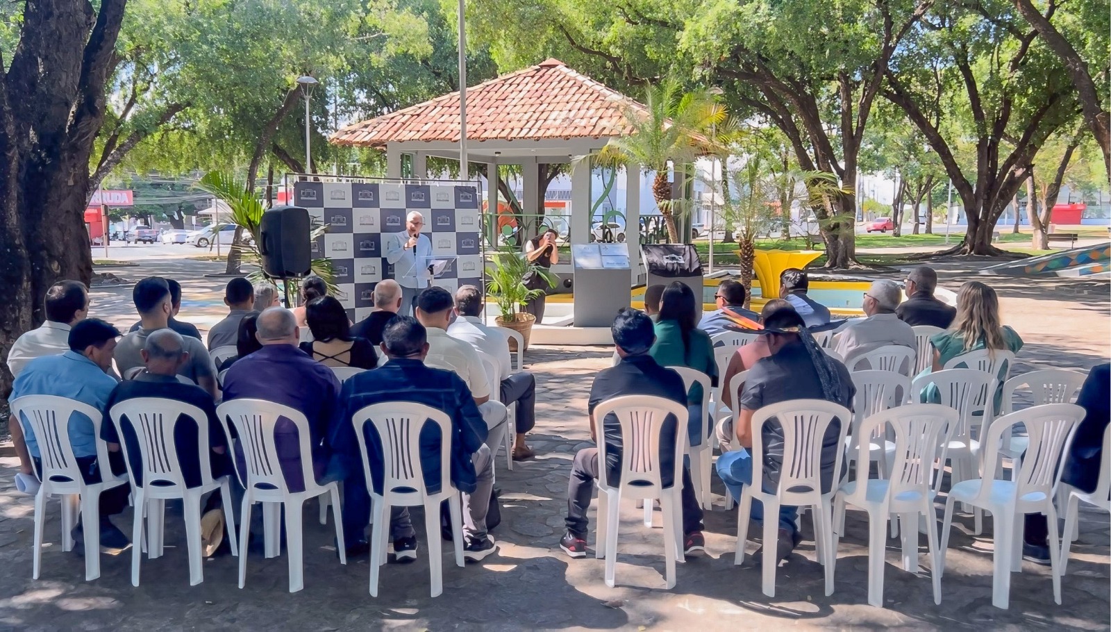 Imagem colorida mostra o desembargador Almiro Padilha, falando no microfone para público na praça do coreto, durante entrega de placa revitalizada do memorial do indígena Ovelário Tames.Abaixo a frase “Homenagem aos Povos Originários - Memorial Indígena revitalizado no Dia dos Direitos Humanos reforça luta por Justiça em Roraima.”