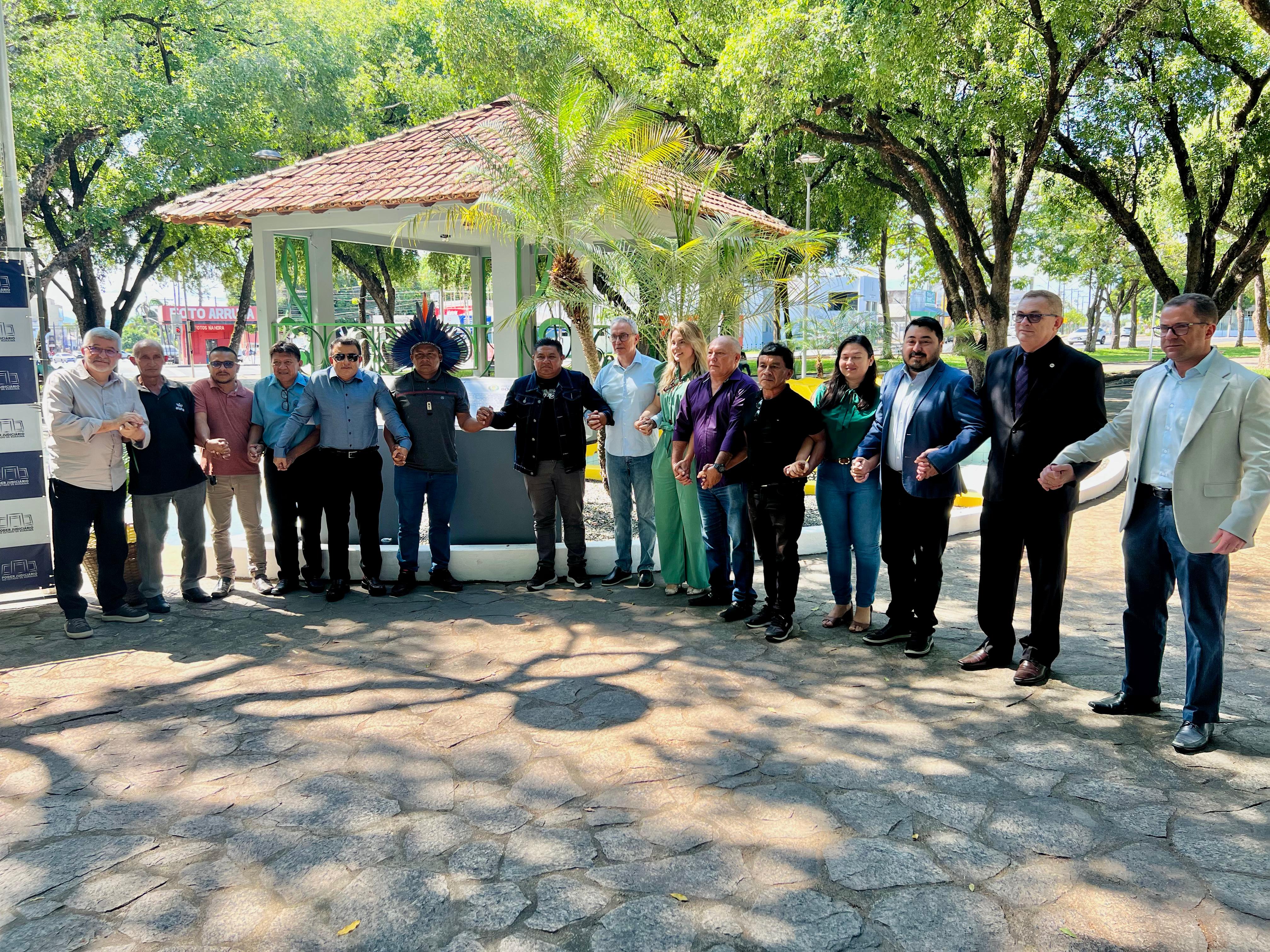 Imagem colorida mostra o desembargador Almiro Padilha, posando para fotografia de mãos dadas juntamente com autoridades convidadas na praça do coreto, ao lado da placa revitalizada do memorial do indígena Ovelário Tames.Abaixo a frase “Homenagem aos Povos Originários - Memorial Indígena revitalizado no Dia dos Direitos Humanos reforça luta por Justiça em Roraima.”
