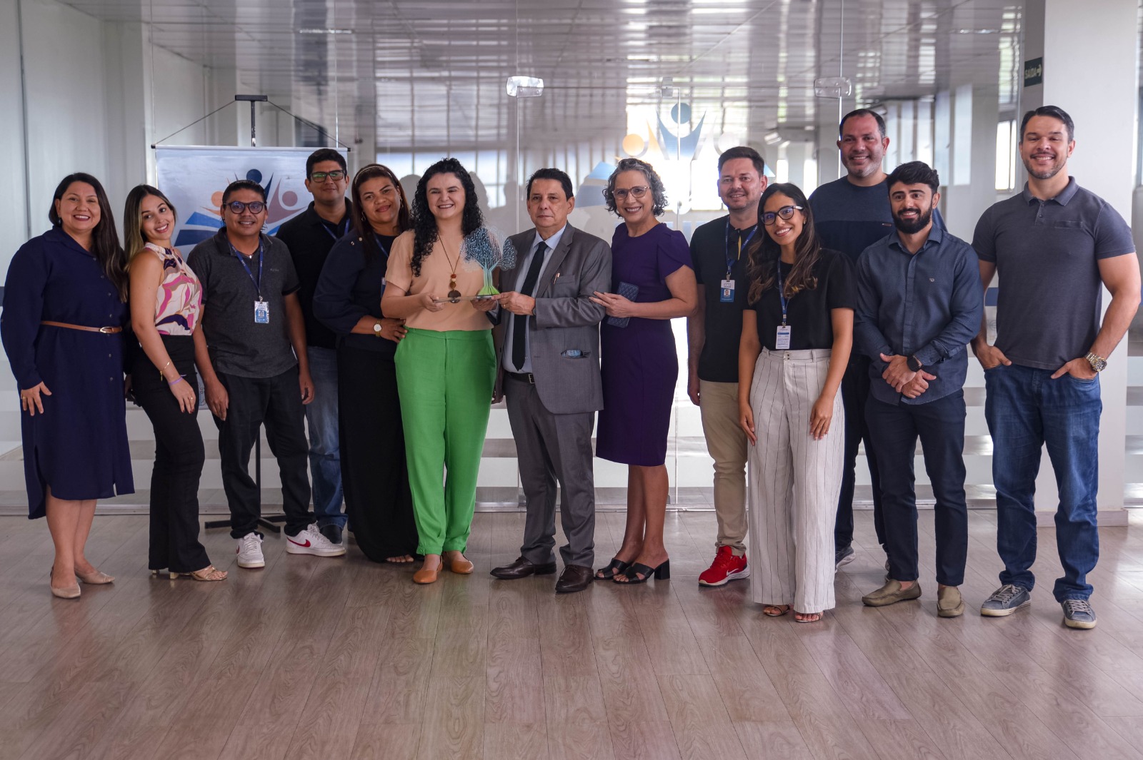 A imagem mostra um grupo de 12 pessoas reunidas em um ambiente corporativo, posando para a foto. Ao centro, o presidente do TJRR, desembargador Jésus Nascimento e a coordenadora acadêmica da Ejurr, Tatiana Saldanha seguram o Prêmio Cnj de qualidade na categoria ExcelÊncia Ao fundo, há um banner e um espaço bem iluminado.