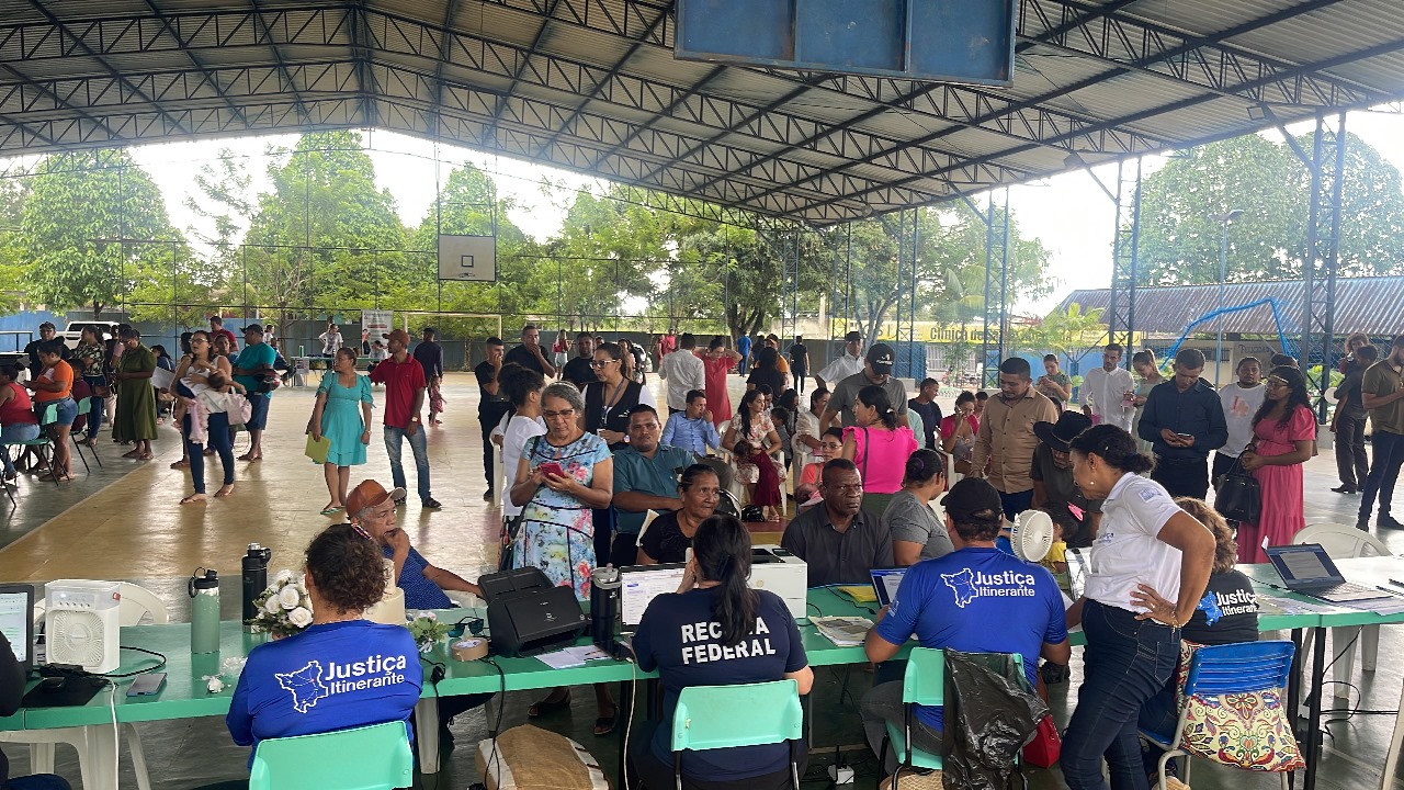 Foto colorida tirada durante atendimentos realizados pela Vara da Justiça Itinerante. A imagem tirada em plano aberto mostra diversas pessoas reunidas em um ginásio de uma escola enquanto esperam para serem atendidas pelos serviços da Justiça Itinerante. 