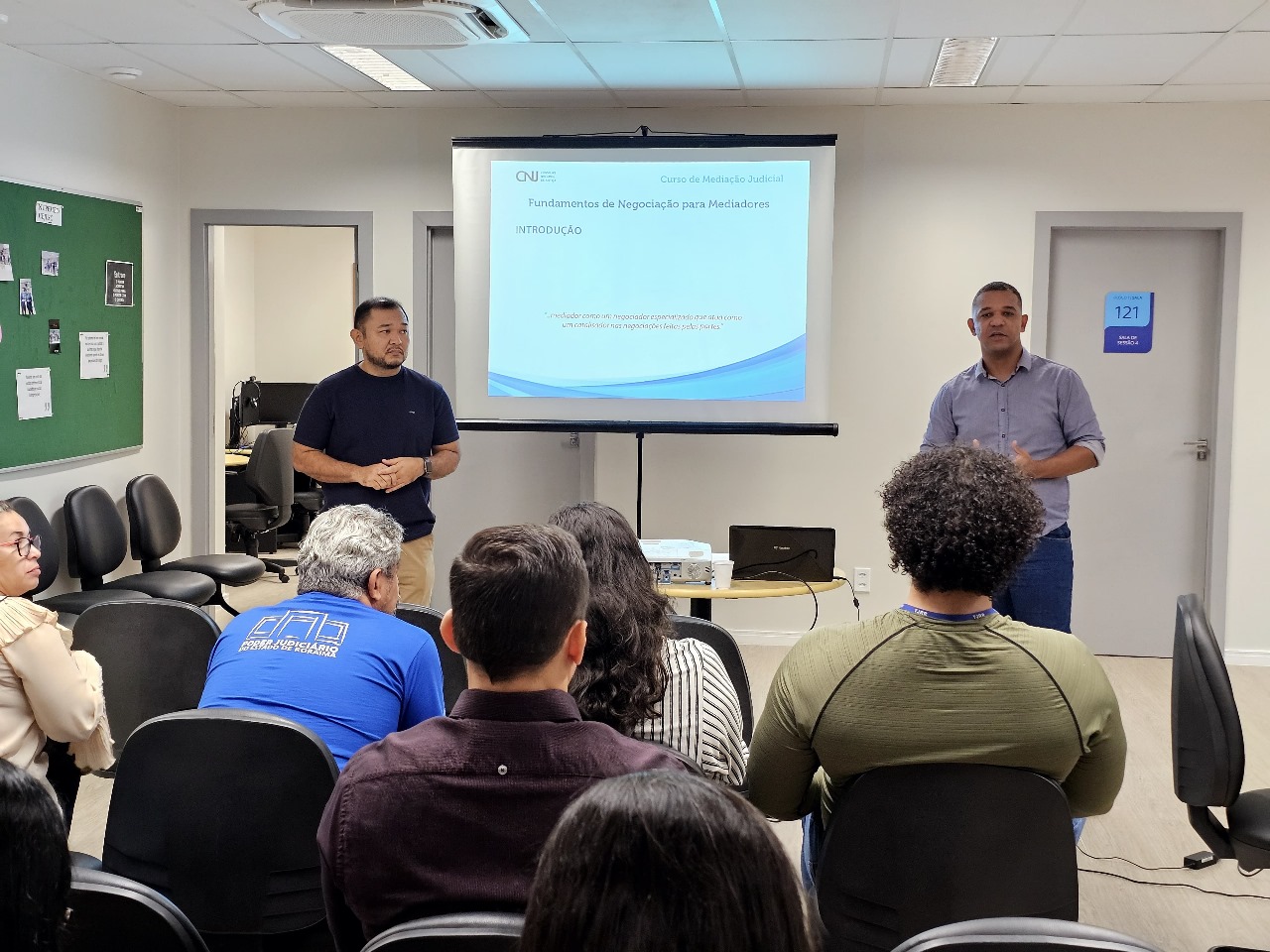 foto colorida mostra uma sala com seis pessoas sentadas prestando atenção na fala de dois palestrantes homens que estão em pé em frente de um slide. 
