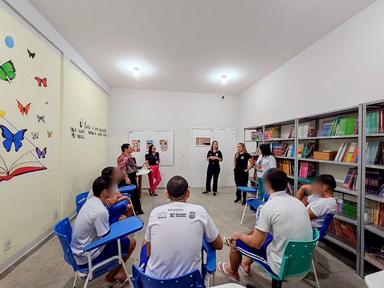 Foto colorida mostra roda de conversa com jovens do CSE Homero de Souza Cruz Filho, durante ação nacional do Conselho Nacional de Justiça (CNJ) em parceria com a editora Cia das Letras, iniciada em 30 de janeiro. A imagem contém dez pessoas, sendo cinco jovens homens e cinco mulheres, entre agentes do Centro e servidoras do projeto Leitura Abre Portas do TJRR.  Do lado esquerdo da sala se encontra livros numa estande e do lado direito  da sala encontra-se figuras ilustrativas de emborrachado.
