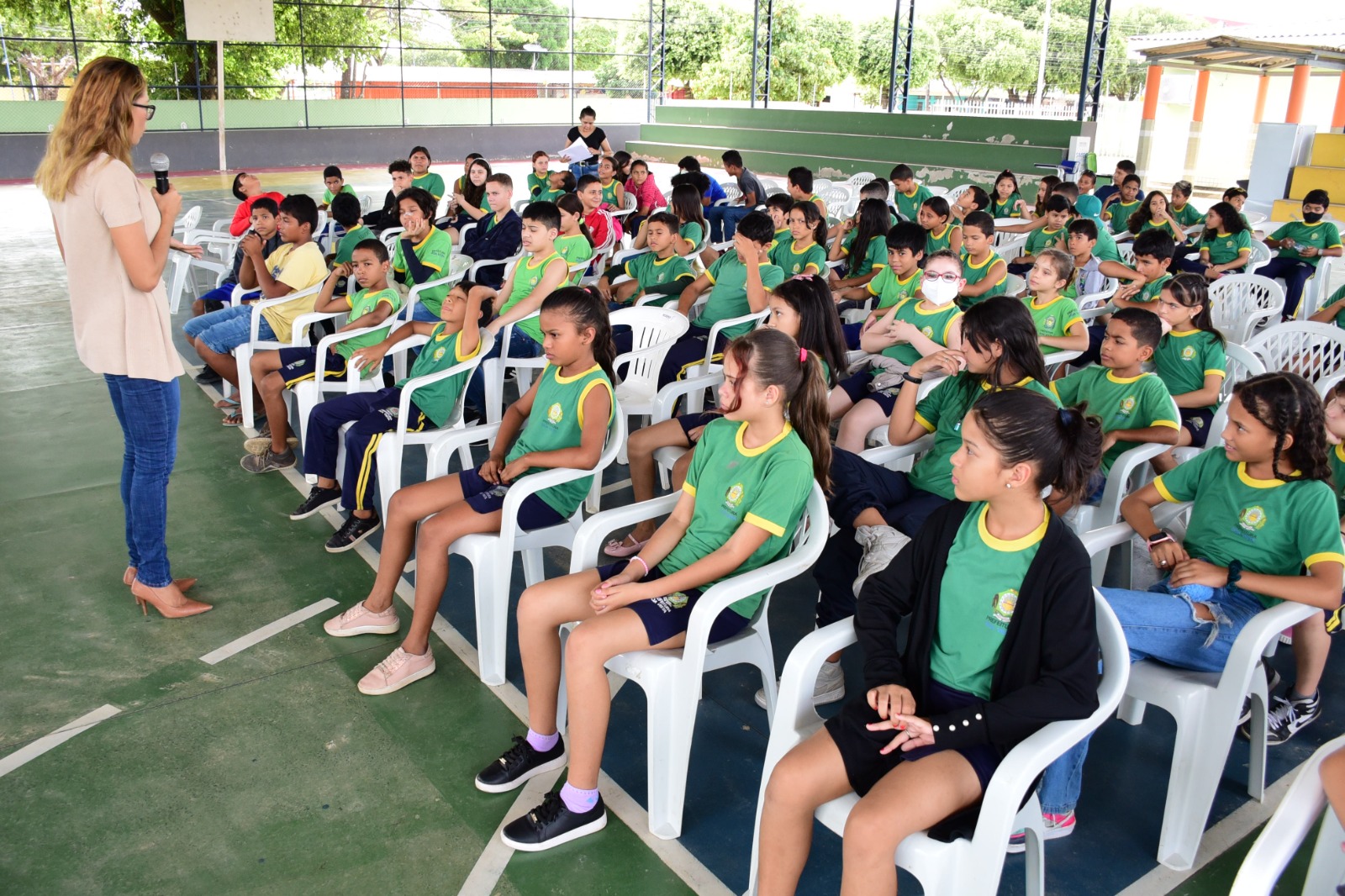 Doméstica e Familiar contra a Mulher Aurilene Mesquita em pé com um microfone na mão. Imagem mostra  mostra os alunos do 5º ano da Escola Municipal Carlos Raimundo sentados em cadeiras brancas no centro de uma quadra de esporte à frente está a chefe do Setor de Atividades de Enfrentamento à Violência 