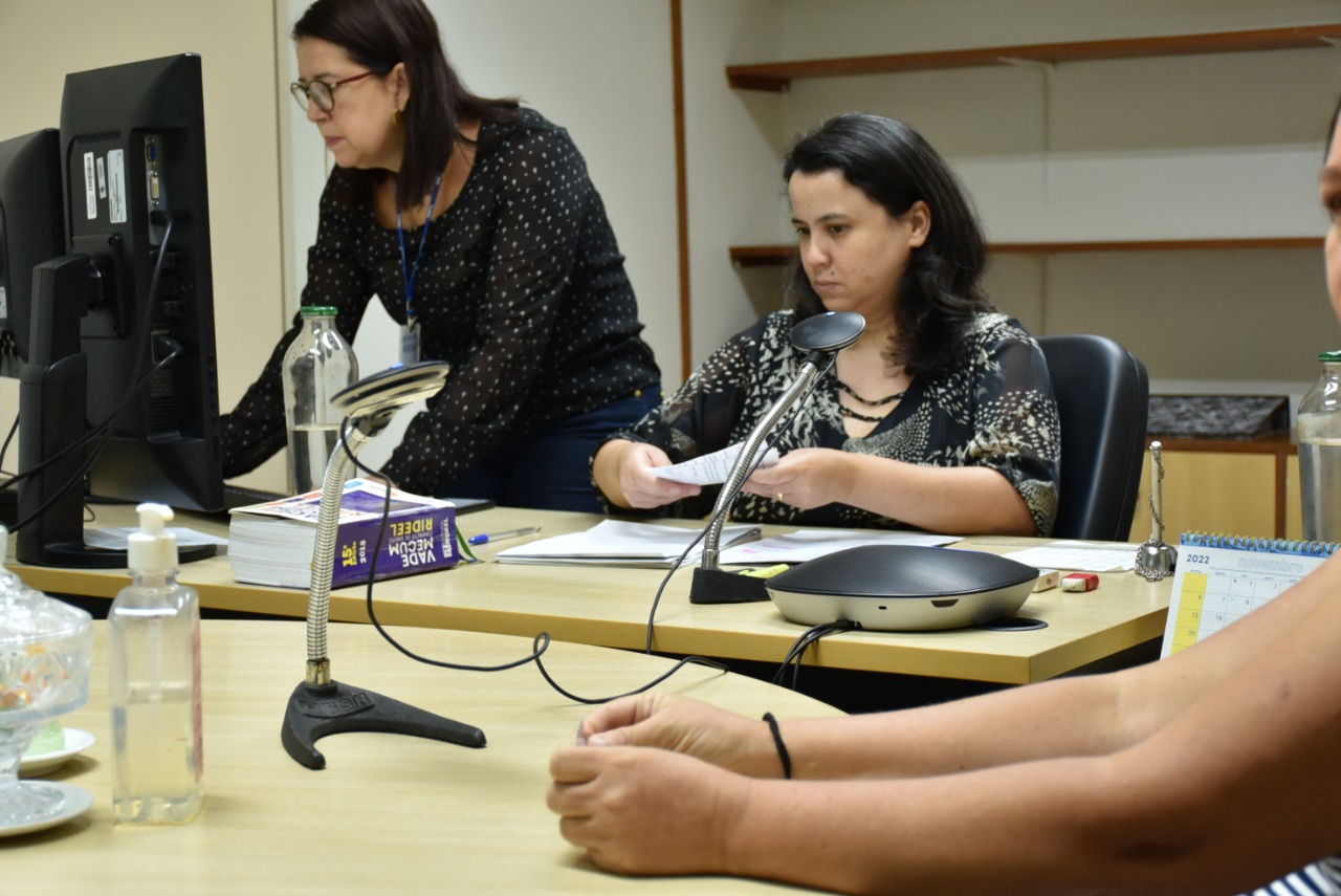 Imagem mostra a juíza titular da 2ª Vara da Família, dra Joana Sarmento, em uma mesa, assessorada por uma servidora, atrás dela, que acessa informações em um computador. A frente dela, aparecem as mãos de uma jurisdicionada atendida pela ação. 