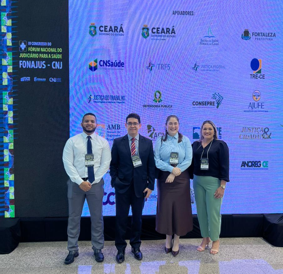  imagem colorida de duiz coordenador do Núcleo de Apoio Técnico do Judiciário (NatJus) e  do Comitê Estadual de Saúde, Breno Coutinho; pela juíza do 1º Núcleo de Justiça 4.0 – Saúde, Rafaella Holanda; pela assessora jurídica do Núcleo de Justiça 4.0, Francisca Rodrigues; e pelo chefe do Setor de Gestão do NatJus, Jhemenson Ferreira pousando para fotografia em frente ao um painel do evento.
