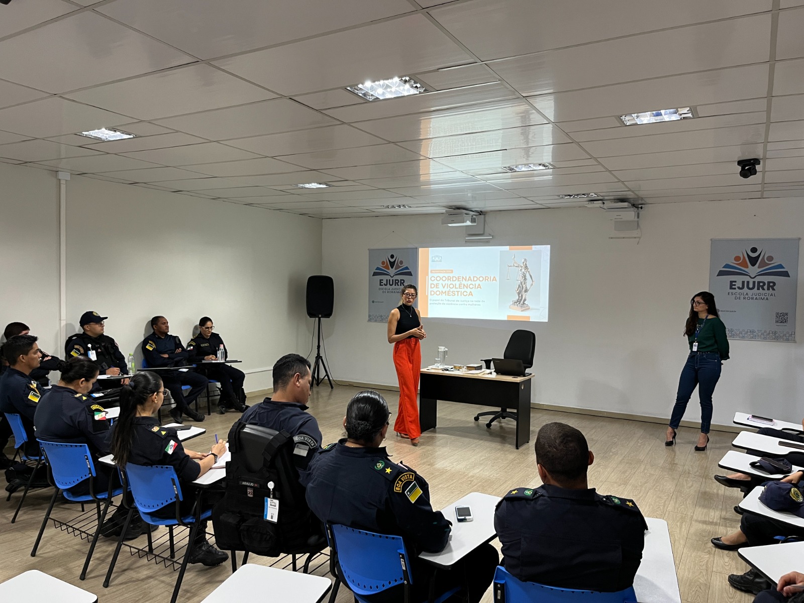 foto colorida de duas servidoras do TJRR a frente da sala e 17 policias sentados em cadeiras escolares