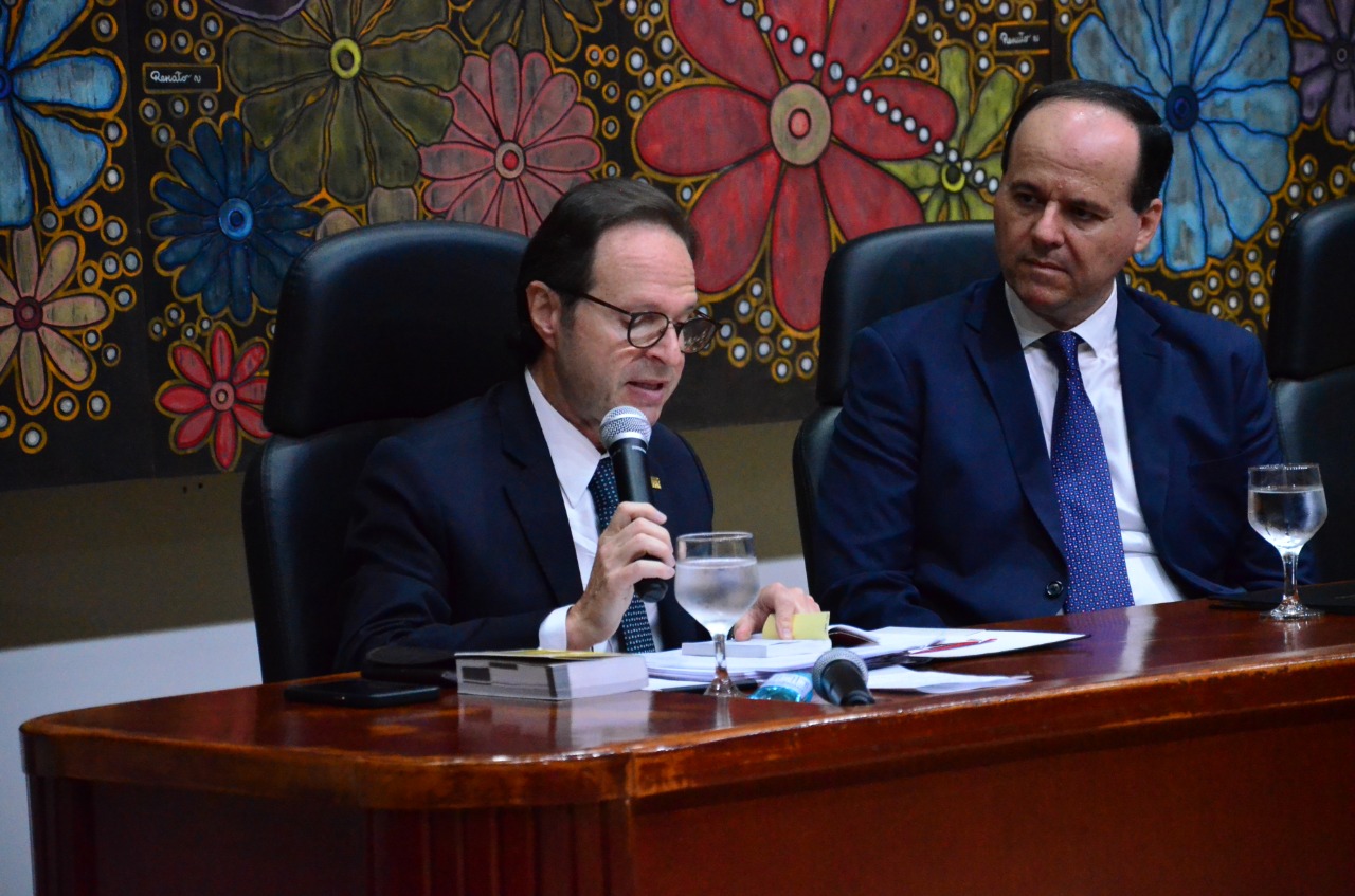 Ministro do STJ segura um microfone e faz uma leitura.  Ele usa óculos.  Ele é observado com atenção pelo presidente do TJRR.  Ambos estão de terno e gravata, sentados atrás de uma mesa de madeira.