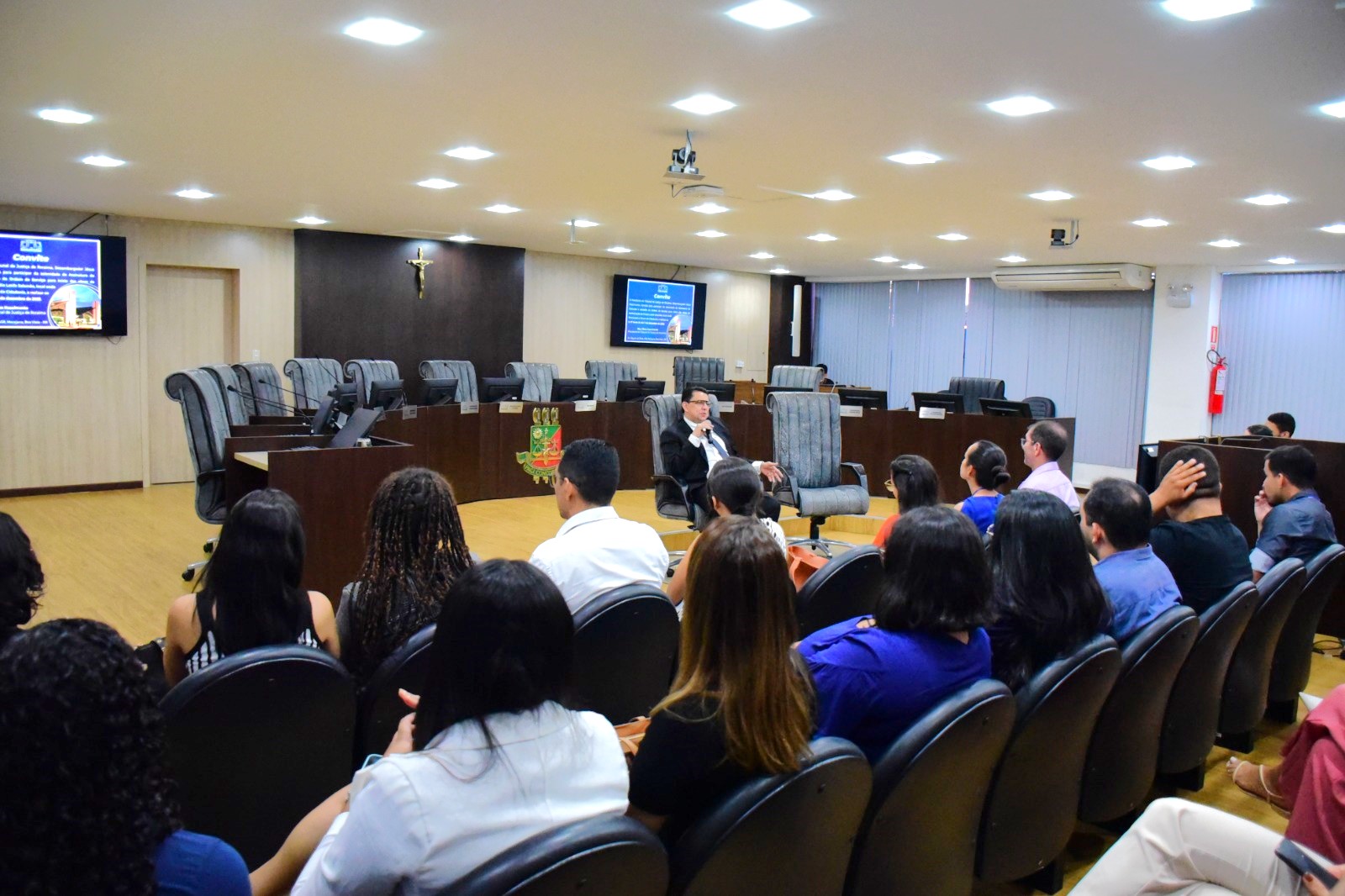 :  fotografia com os residentes estão sentados em direção ao presidente do TJRR, desembargador Jésus Nascimento que está sentando falando ao microfone