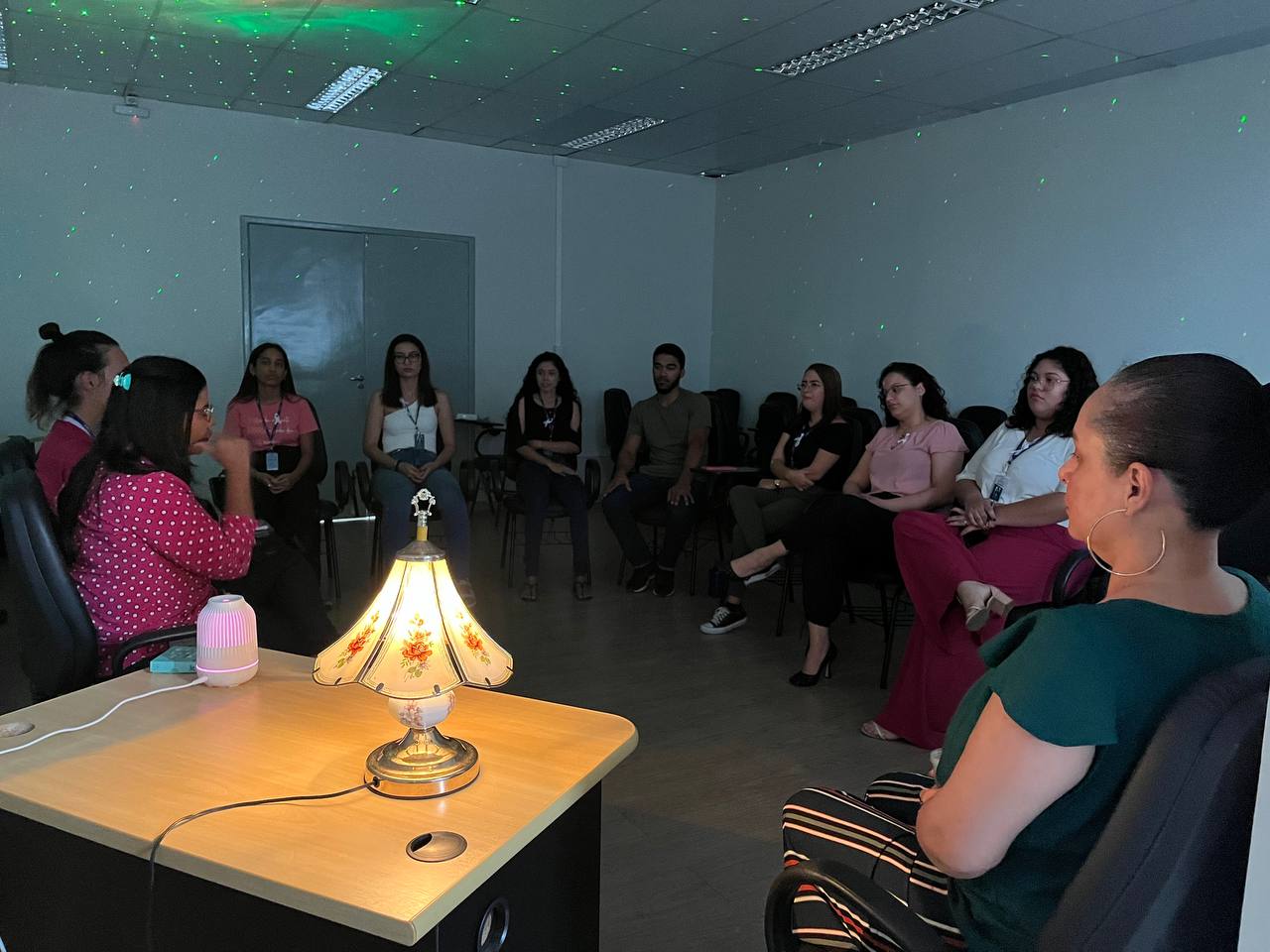 foto colorida de pessoas em uma sala meditando. O ambiente esta com pouca iluminação 