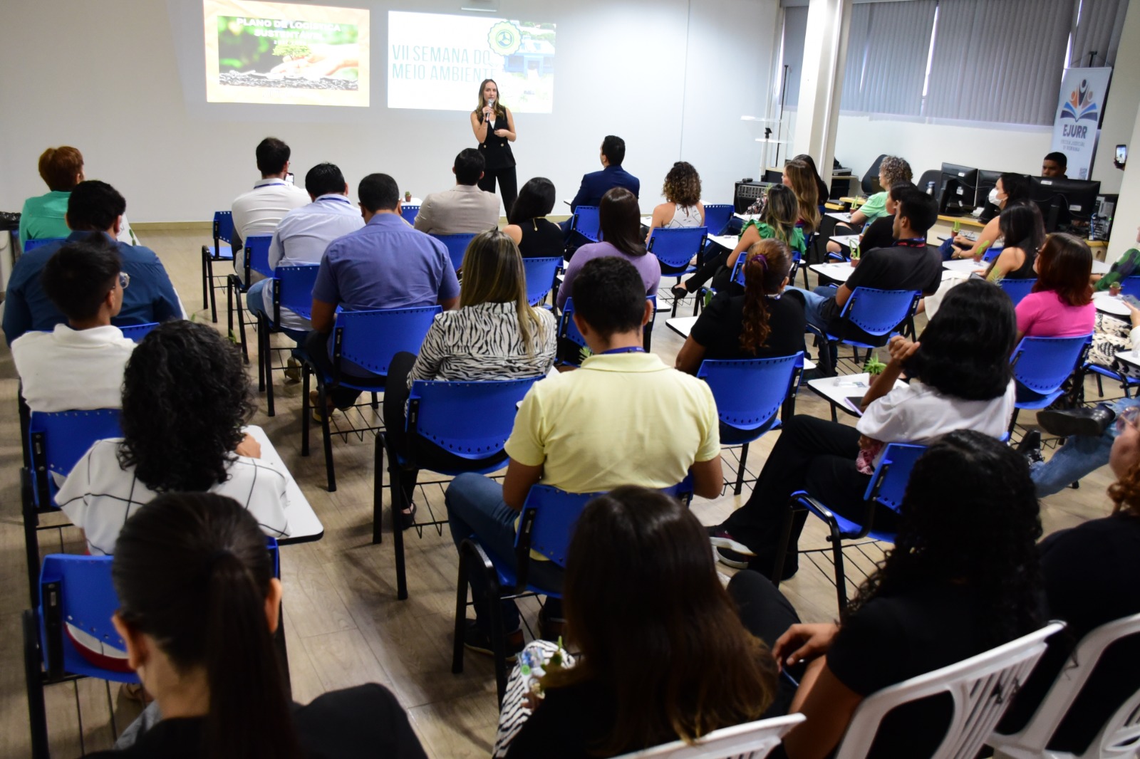 Foto colorida mostra servidores e magistrados sentados de costas para a foto, acompanhando a fala da chefe do Setor de Gestão Socioambiental e Acessibilidade, Ana Livia Sá, que está em pé, frente a platéia, discursando ao microfone. 