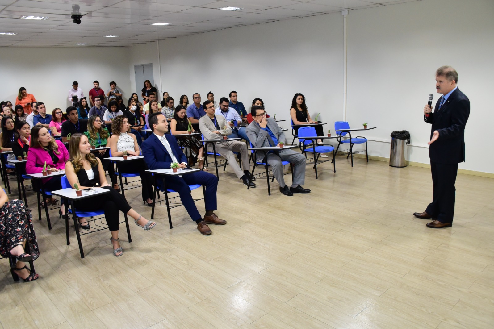 foto colorida mostra o presidente da Comissão Gestora do Plano de Logística Sustentável do TJRR, juiz Jarbas Lacerda de Miranda, a direita em pé, falando ao microfone na solenidade de abertura da 7ª Semana do Meio Ambiente. Mais à esquerda estão cerca de 50 pessoas, sentadas em cadeiras azuis, acompanhando a fala do presidente. 