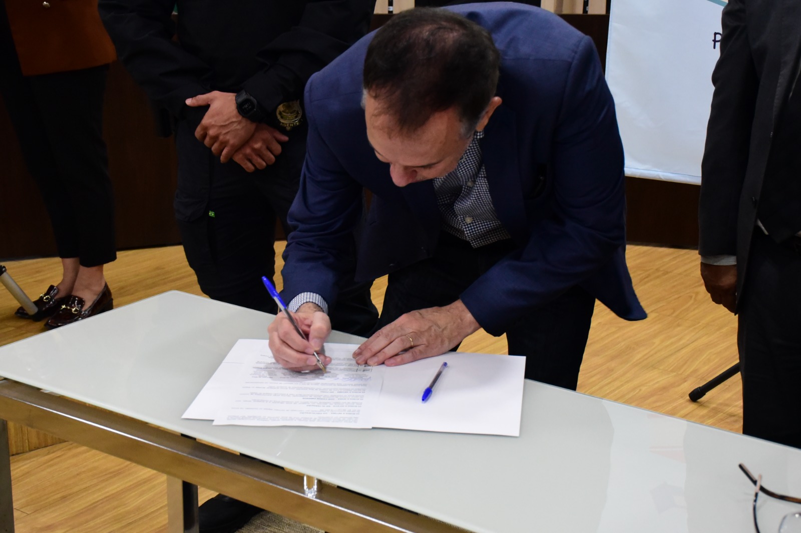 foto colorida dando enfoque no  desembargador, supervisor do GMF, Almiro Padilha, assinando o acordo de cooperação entre o TJRR, Receita Federal e Sejuc que está em cima de uma mesa de vidro branco com um caneta bic azul. 