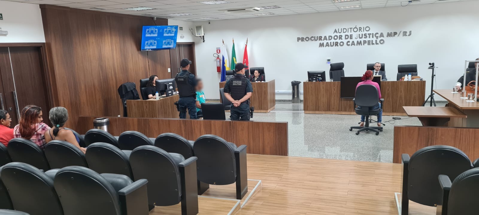 Imagem mostra o tribuna do júri no momento da oitiva de uma testemunha, sentada em uma cadeira em frente ao juiz. A imagem mostra o réu sentado ao lado, escoltado por dois policiais, além de três mulheres sentadas na plateia