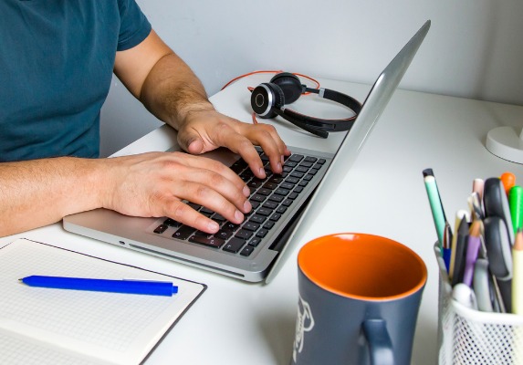 Foto de mãos de pessoa mexendo em um notebook que está sobre uma mesa, que também têm um fone, uma xícara, um papel e várias canetas sobre ela.