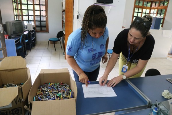 Servidora do Tribunal de Justiça e servidora da Prefeitura de Boa Vista, olham papel que está sobre uma mesa, que também tem duas caixas cheias de pilhas em cima dela.