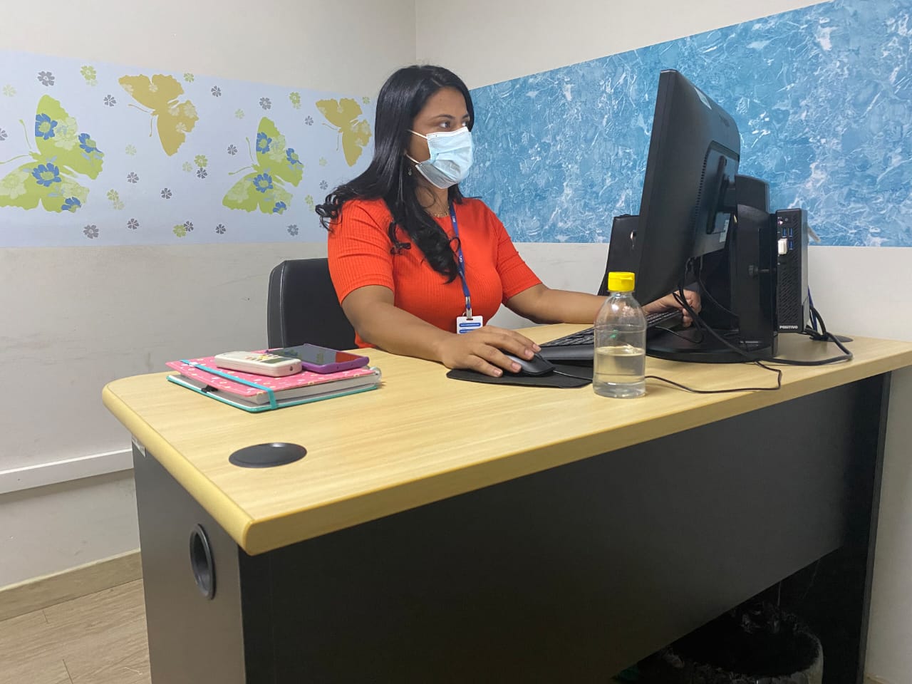 Servidora do tribunal de Justiça sentada atrás de uma mesa, mexendo no computador em uma sala com dois quadros na parede.