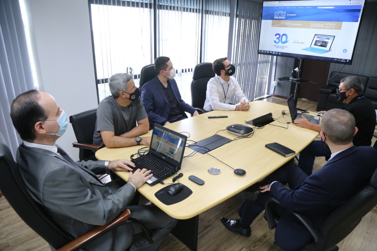 Presidente do Tribunal de Justiça, desembargador Cristóvão Suter sentado em mesa de reuniões com servidores e um notebook em frente à ele, olhando uma TV que apresenta o visual do novo portal.