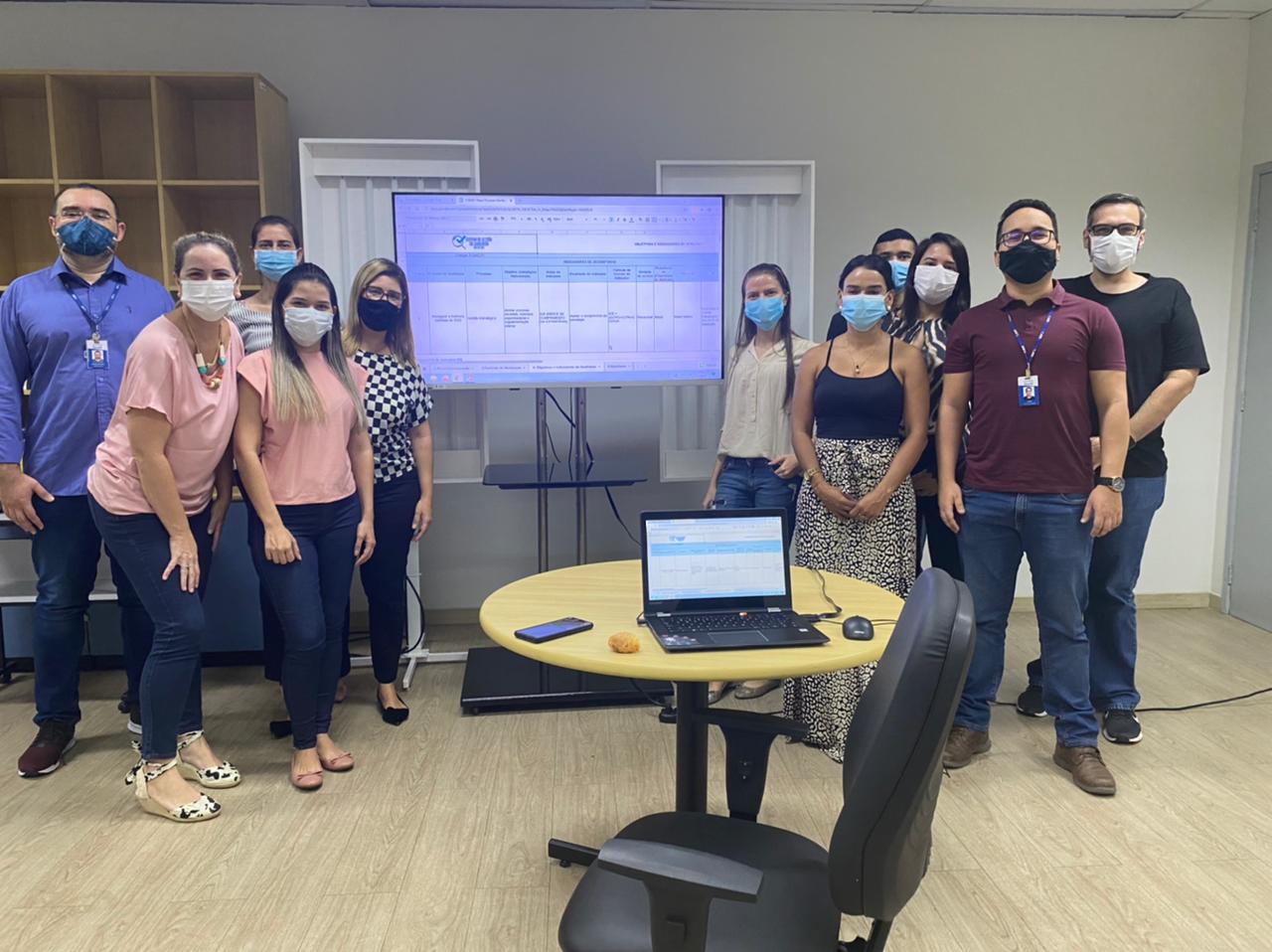 Servidores da Secretaria de Gestão Estratégica posam para foto, ao lado de uma TV e com uma cadeira, e uma mesa com um computador em cima.