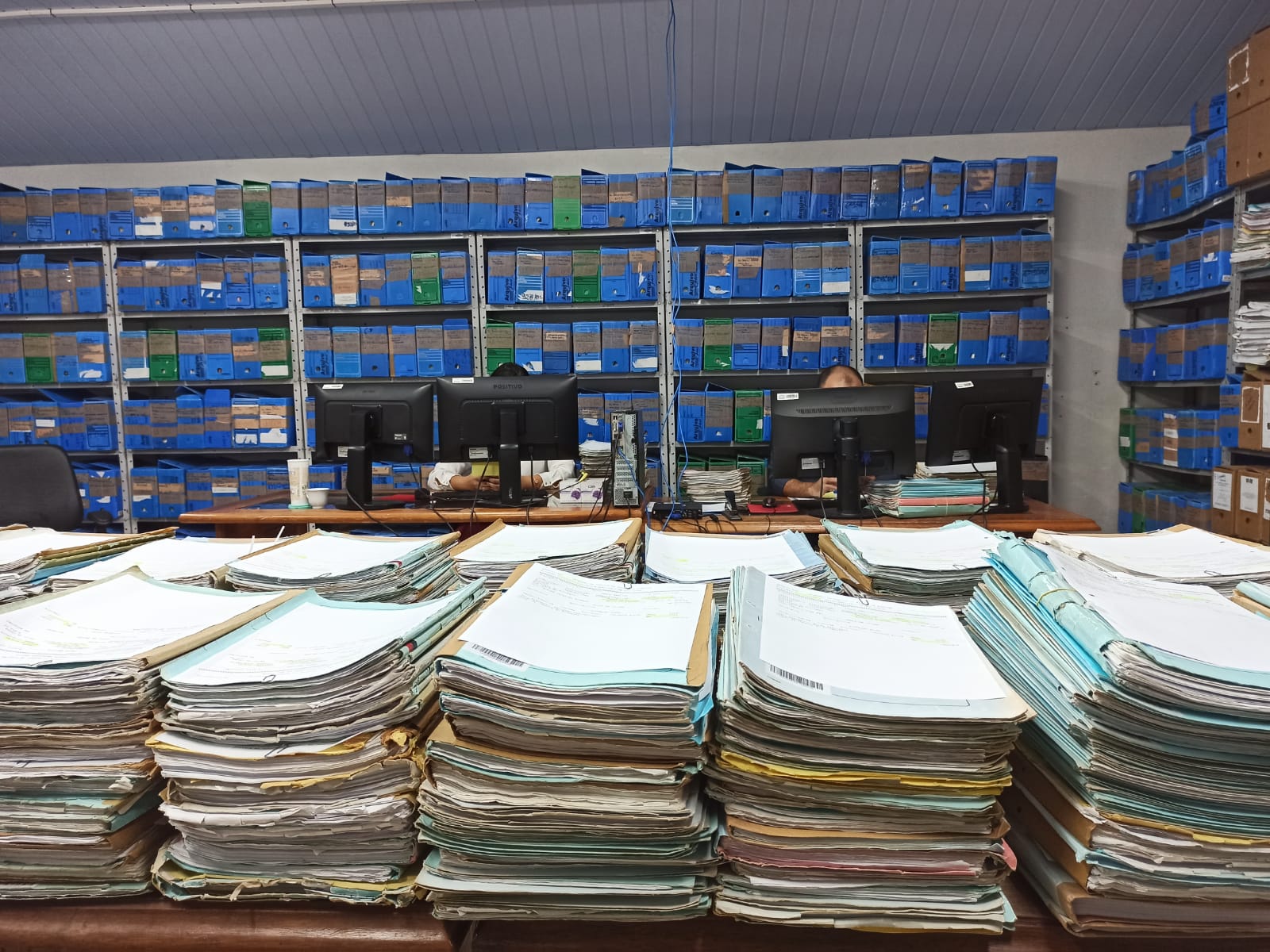 Fotos de documentos empilhados em cima de uma mesa em primeiro plano; pessoas atrás de computadores e armários com arquivos ao fundo no Setor de Arquivo da Subsecretaria de Gestão Documental do Tribunal de Justiça de Roraima.