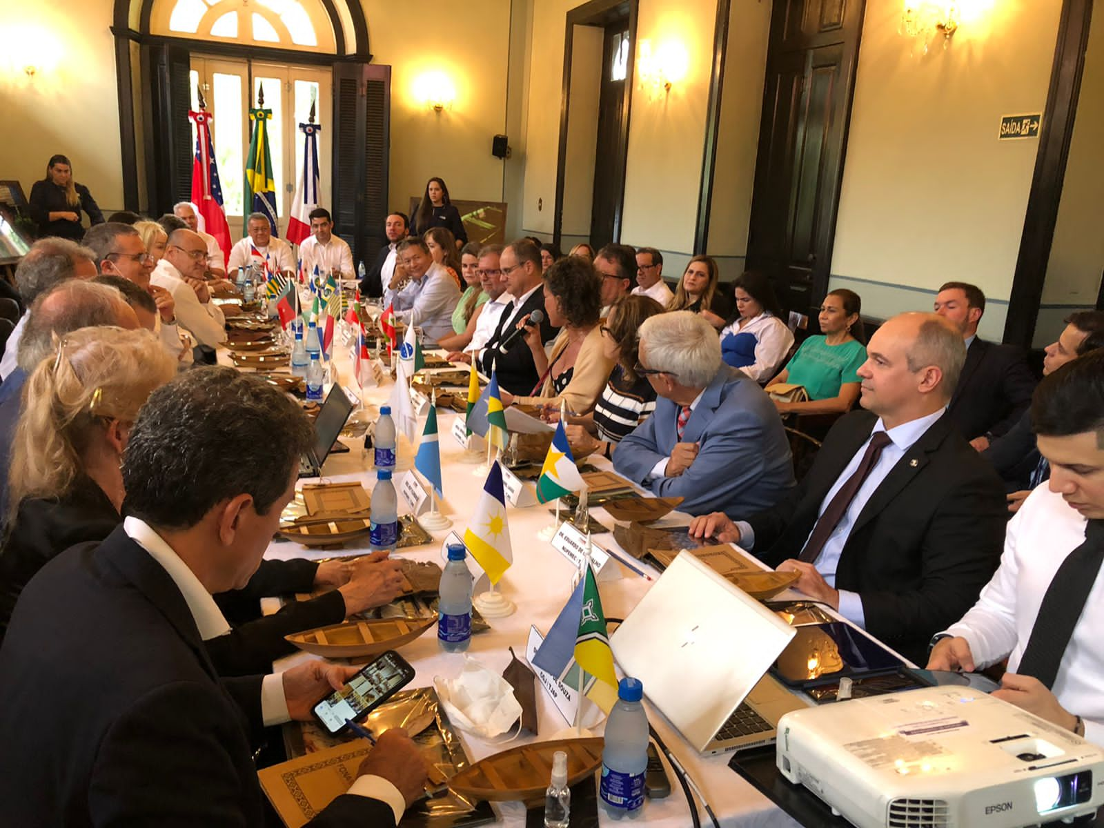 Imagem colorida mostra uma mesa com pequenos artesanatos de madeira em forma de canoas, garrafas de água e  pequenas bandeiras hasteadas de todos os Estados Brasileiros e ao redor estão sentados os representantes de cada Estado.  