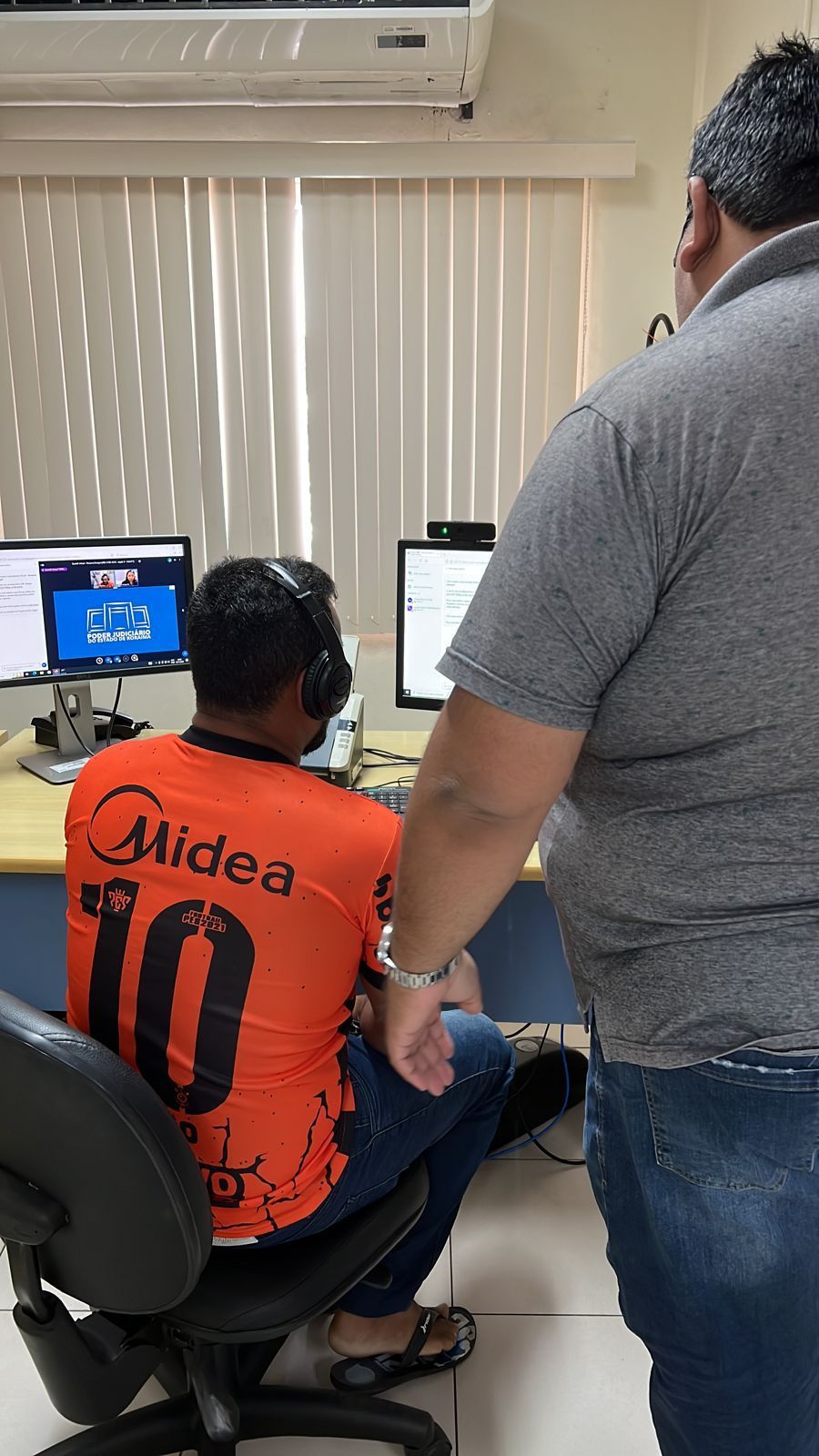 Imagem colorida mostra dois homens, um deles em pé e outro sentado com um fone de ouvido, ambos estão de costas olhando para a tela de um computador posta sobre uma mesa.