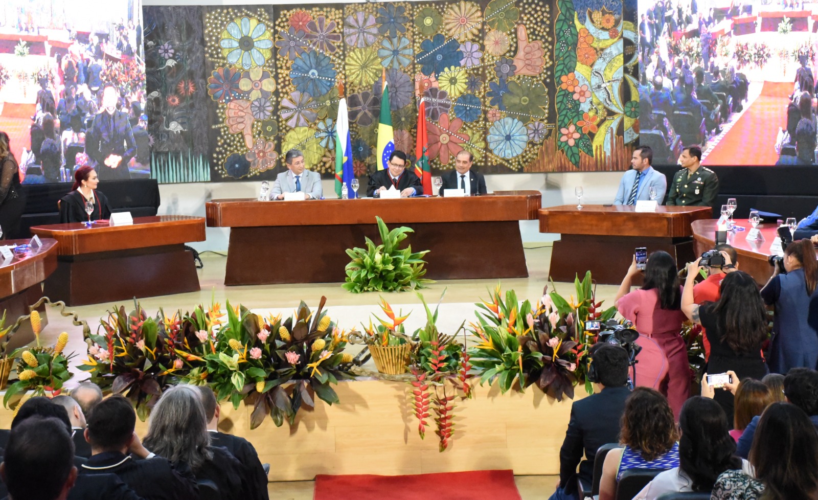 Imagem colorida em formato retangular mostra a parte interna do auditório do Fórum Cível Advogado Sobral Pinto.  A frente está a mesa diretora do tribunal de Justiça.  Acima, posicionado de forma centralizada está um quadro mosaico com flores coloridas e cada lado há telões exibindo uma cerimônia que ocorre no local.