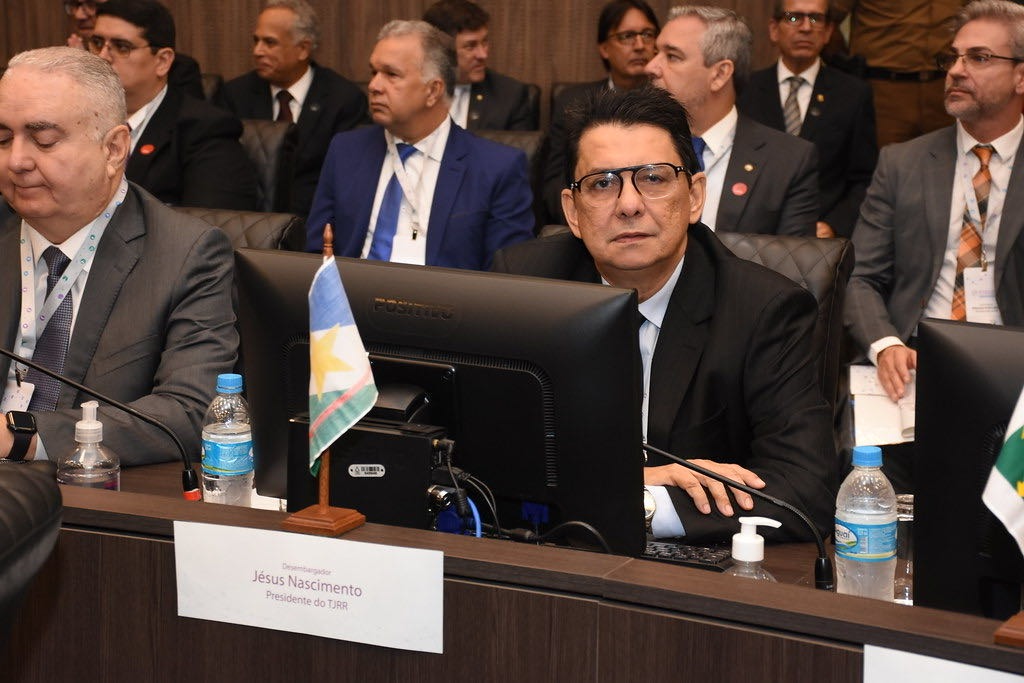 Imagem mostra o presidente do Tribunal de Justiça de Roraima sentado em frente a um computador, olhando para a câmera, com outros presidentes ao seu lado e atrás dele