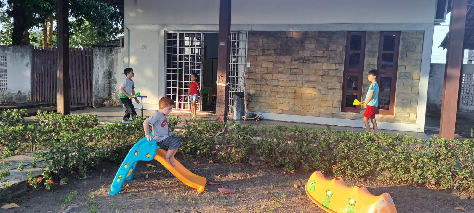 Imagem colorida mostra uma área externa arborizada. No local há quatro crianças brincando.