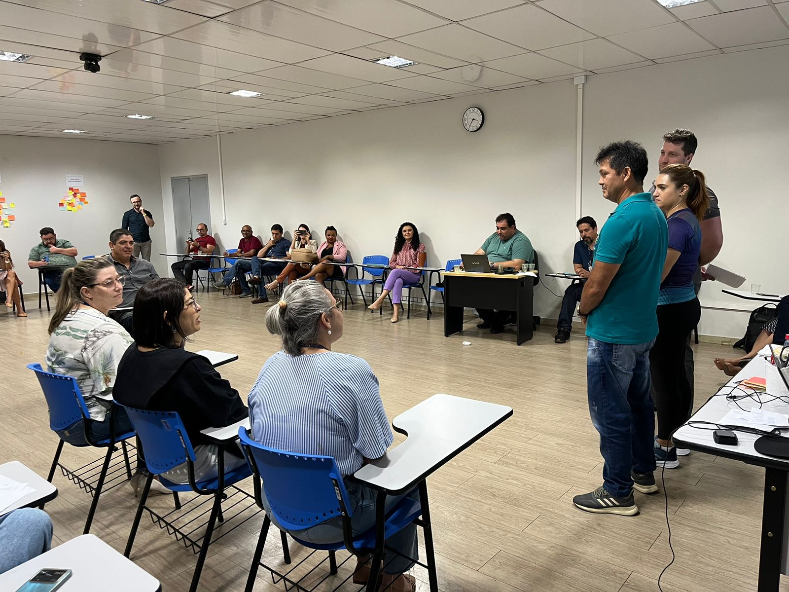 foto colorida de servidores sentados em cadeiras escolares, em meio circulo enquanto observam outros dois em pé na rente apresentando 