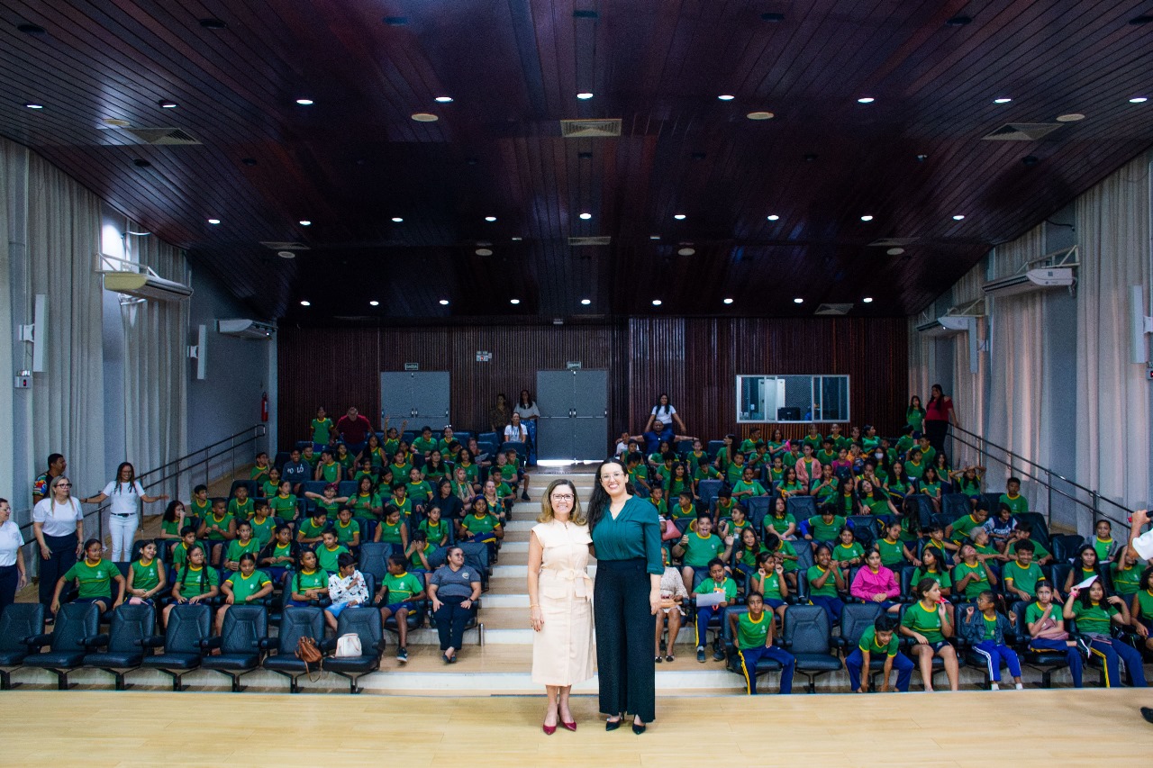 Auditório cheio de estudantes uniformizados em verde, acompanhando uma atividade. Duas mulheres estão em destaque na frente do palco, entre elas a juiza Suelen Alves, posando para a foto. O ambiente é bem iluminado, com teto de madeira e cadeiras organizadas em fileiras.