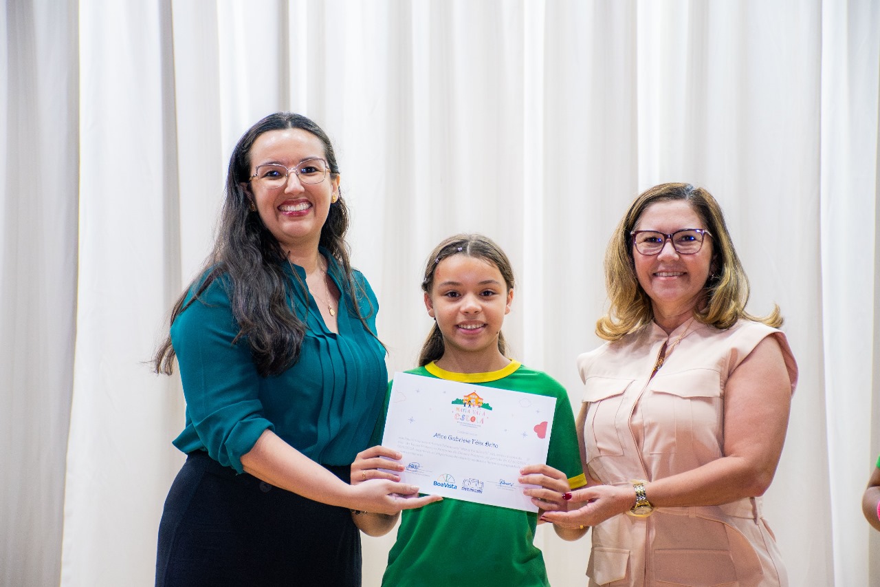 Foto colorida mostrando a juíza Suellen Alves, ao lado da secretária municipal de Educação, Maria Consuêlo Sales Silva, e de uma estudante enquanto ela segura o certificado do projeto "Maria Vai à Escola".