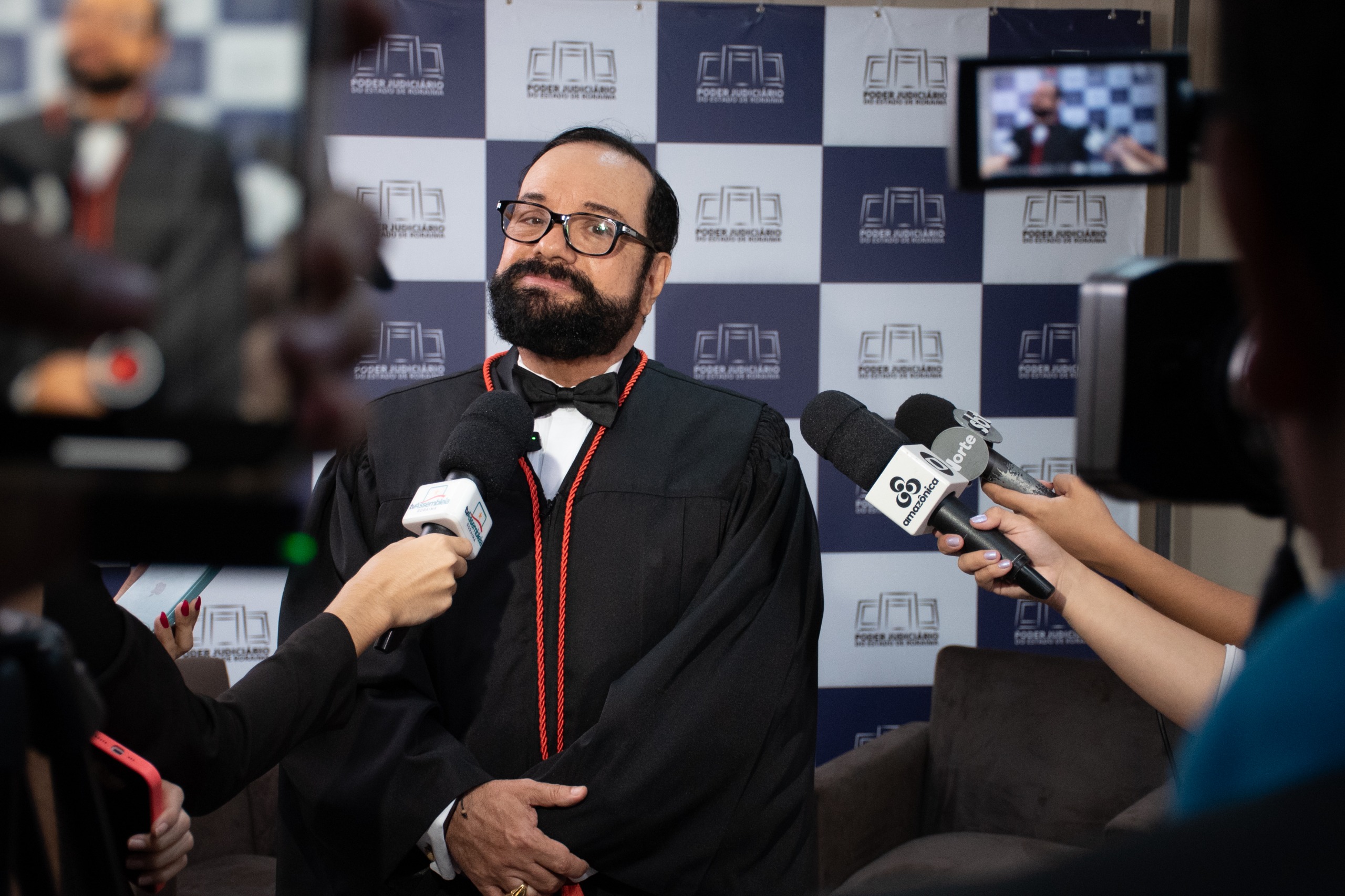 foto colorida mostra o desembargador Leonardo Cupello trajando a vestimenta toga sendo entrevistado por jornalistas. Ele está diante de um fundo com o logotipo do Poder Judiciário do Estado de Roraima. Diversos microfones e câmeras estão apontados para ele, durante a coletiva de imprensa.