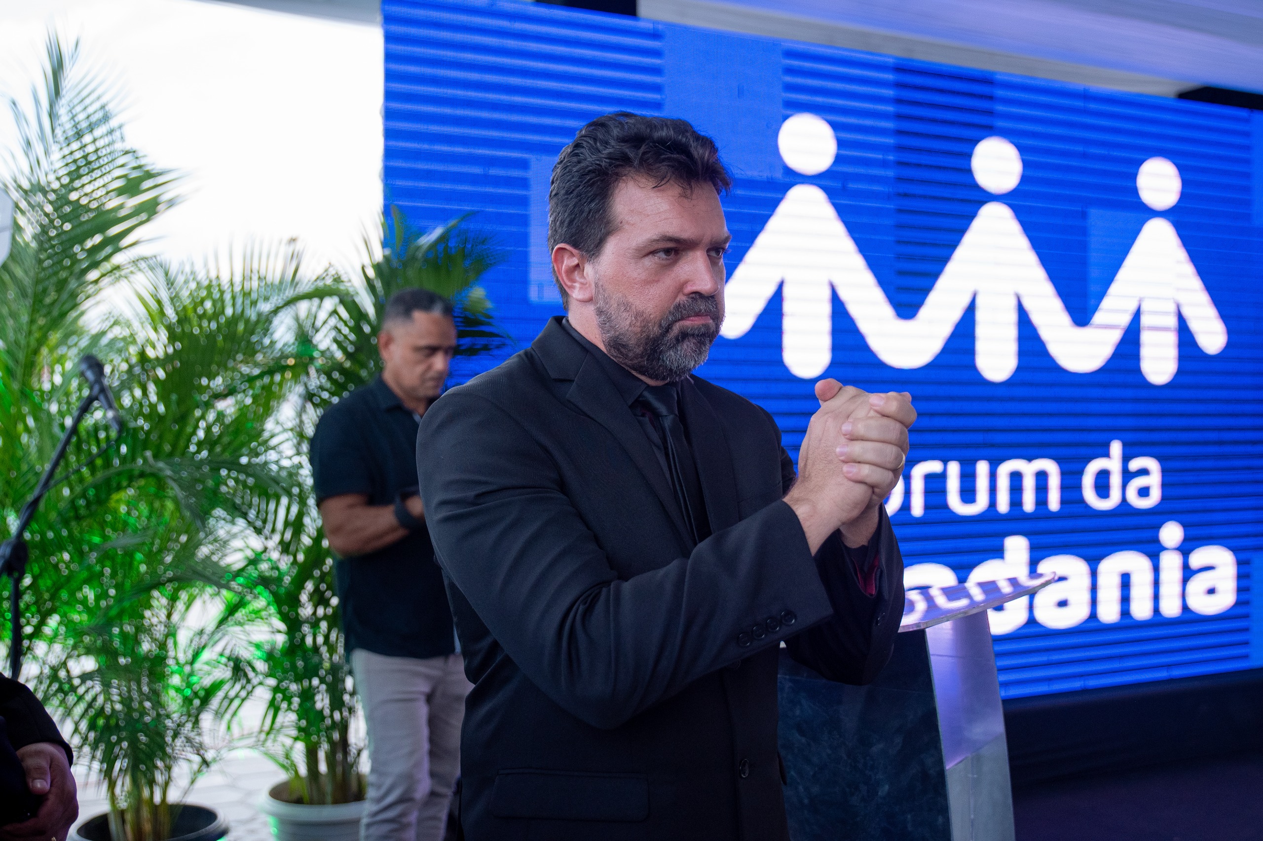 A imagem mostra um homem de terno preto em um evento formal, realizando sinais em Libras, pois ele é intérprete de Língua Brasileira de Sinais. Ao fundo, há um painel azul com o texto "Fórum da Cidadania" e um símbolo de pessoas unidas, além de plantas e um homem ao fundo usando roupa preta.