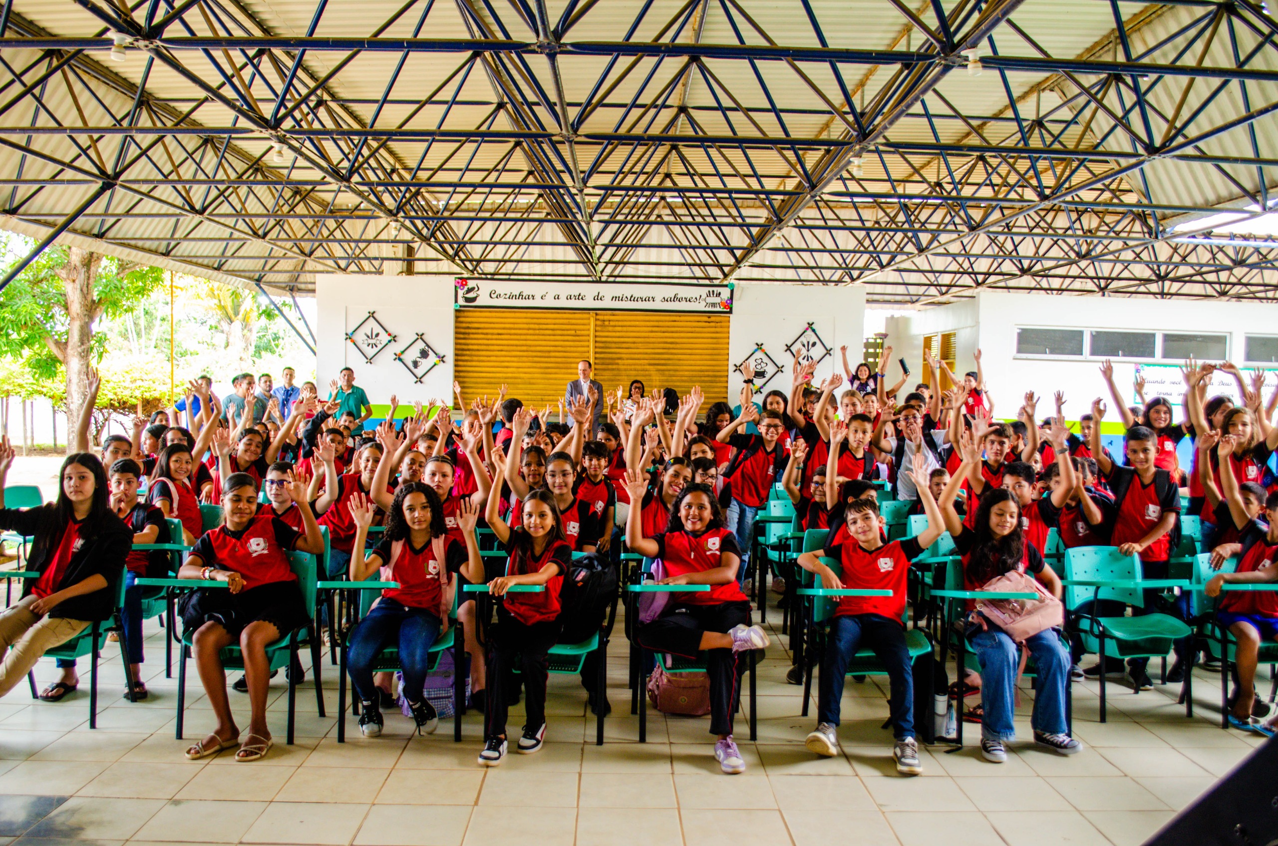 Imagem colorida contém um grande grupo de estudantes de uniforme vermelho sentados em cadeiras verdes, sorrindo e levantando as mãos em um ambiente coberto e iluminado.