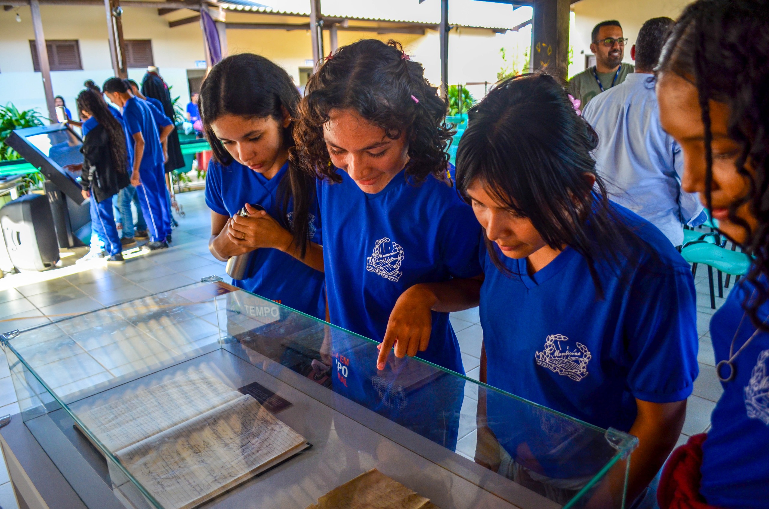 Imagem colorida contém um grupo de estudantes de uniforme azul observando atentamente documentos antigos expostos em uma vitrine de vidro, em um ambiente iluminado e movimentado.