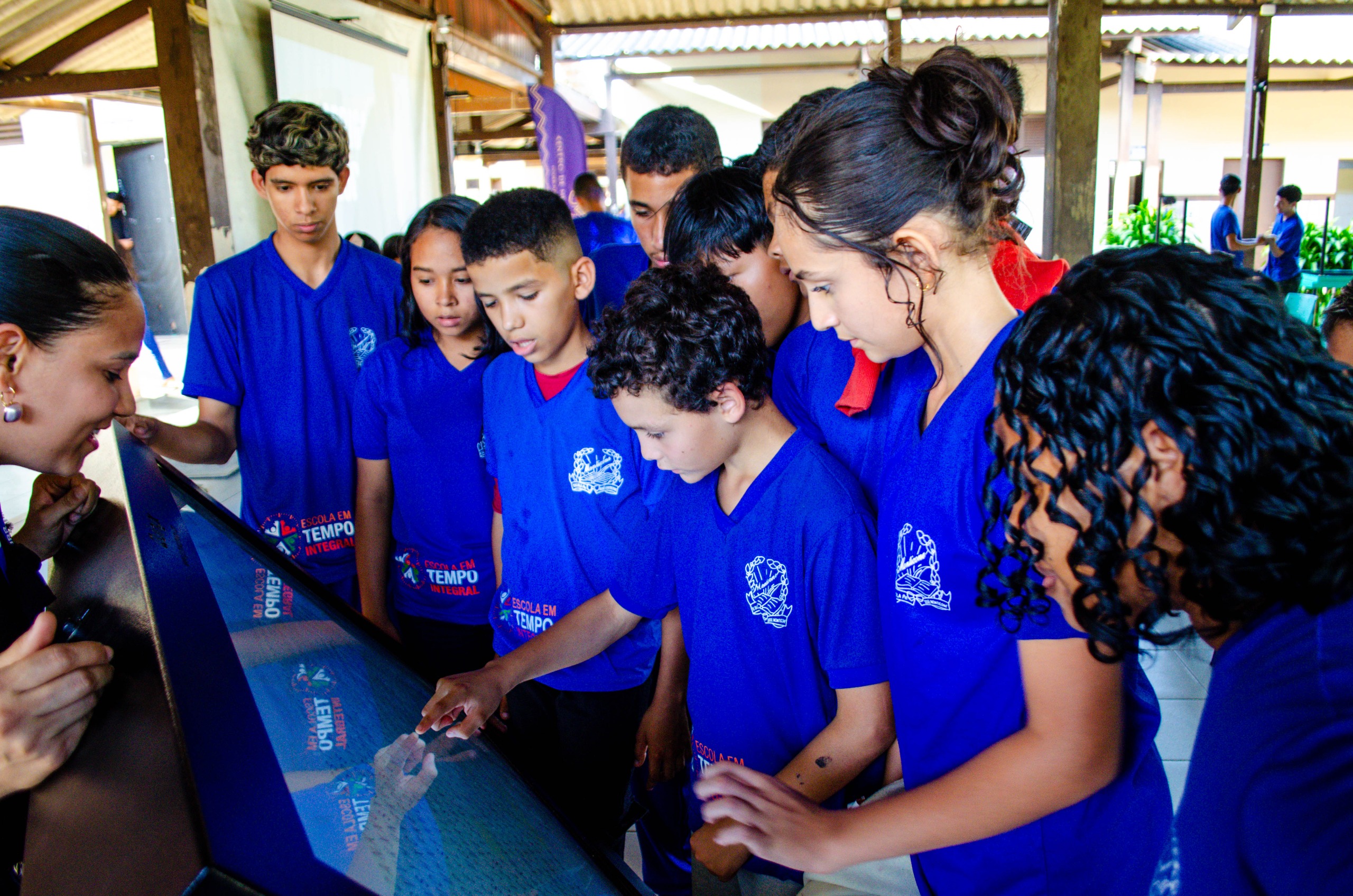 Imagem colorida contém um grupo de estudantes de uniforme azul reunidos em torno de uma tela interativa, tocando e observando atentamente enquanto uma mulher os orienta.