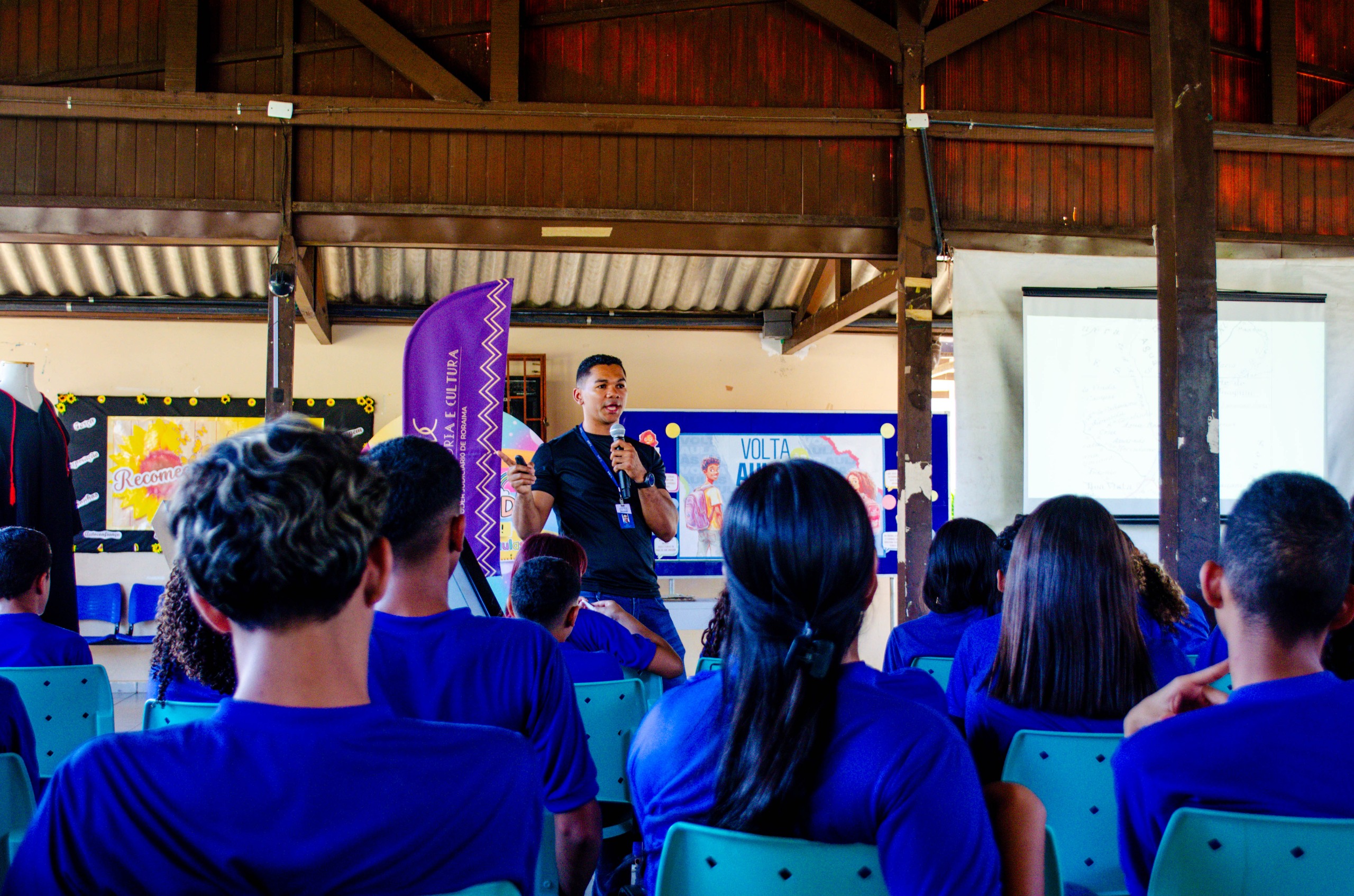 Imagem colorida contém um grupo de estudantes de uniforme azul, sentados em carteiras verdes e de costas para a câmera, prestando atenção enquanto um homem discursa.