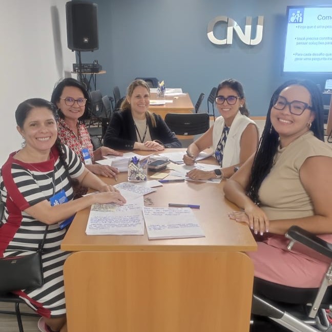 Imagem colorida mostra participantes em um evento no Conselho Nacional de Justiça (CNJ), reunidos em uma mesa de trabalho com anotações e materiais de estudo. O grupo é composto por mulheres que interagem, escrevem e sorriem para a câmera, entre elas a Desembargadora Tânia Vasconcellos e a servidora Claudete Pereira. Ao fundo, é possível ver o logotipo do CNJ na parede e uma tela com uma apresentação. Imagem colorida mostra participantes em um evento no Conselho Nacional de Justiça (CNJ), reunidos em uma mesa de trabalho com anotações e materiais de estudo. O grupo é composto por mulheres que interagem, escrevem e sorriem para a câmera, entre elas a Desembargadora Tânia Vasconcellos e a servidora Claudete Pereira. Ao fundo, é possível ver o logotipo do CNJ na parede e uma tela com uma apresentação.
