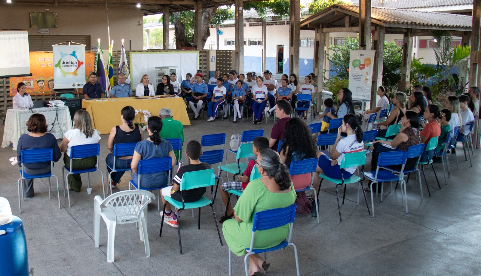 foto colorida de um pátio escolar com pessoas sentadas e a frente uma mesa com servidores 