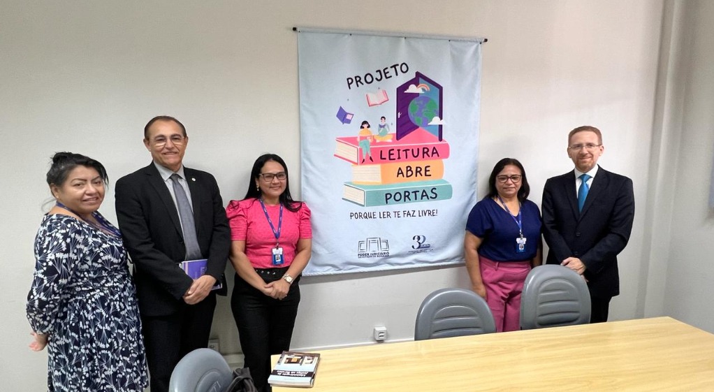 foto colorida de tres servidoras mulheres pousando para a fotografia ao lado dos juízes enfrente ao painel 'leitura abre portas" 