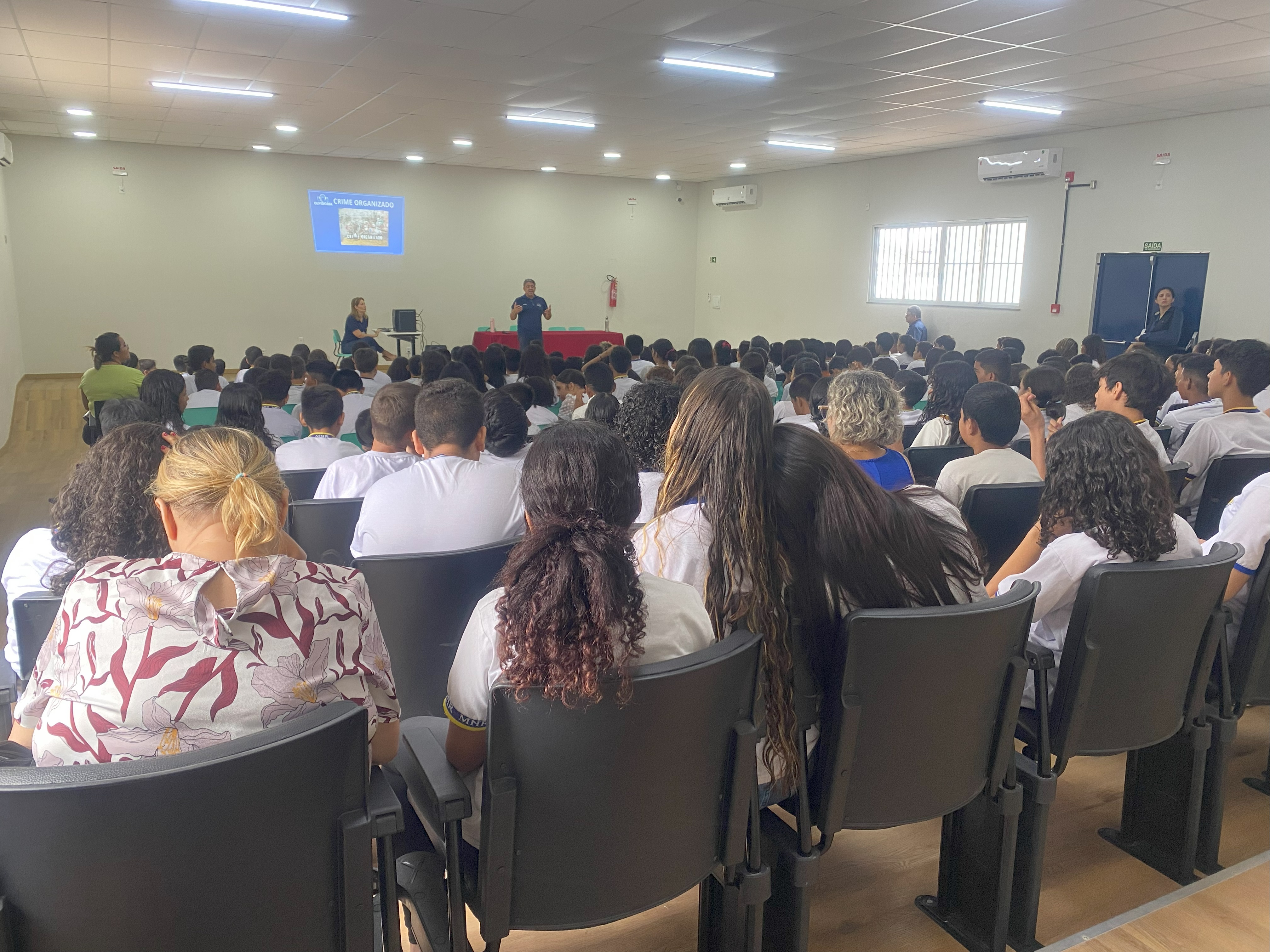 Imagem colorida mostra jovens sentados nas cadeiras do auditório, enquanto o servidor da Ouvidoria-Geral do Tribunal de Justiça de Roraima, Orib Ziedson palestra.